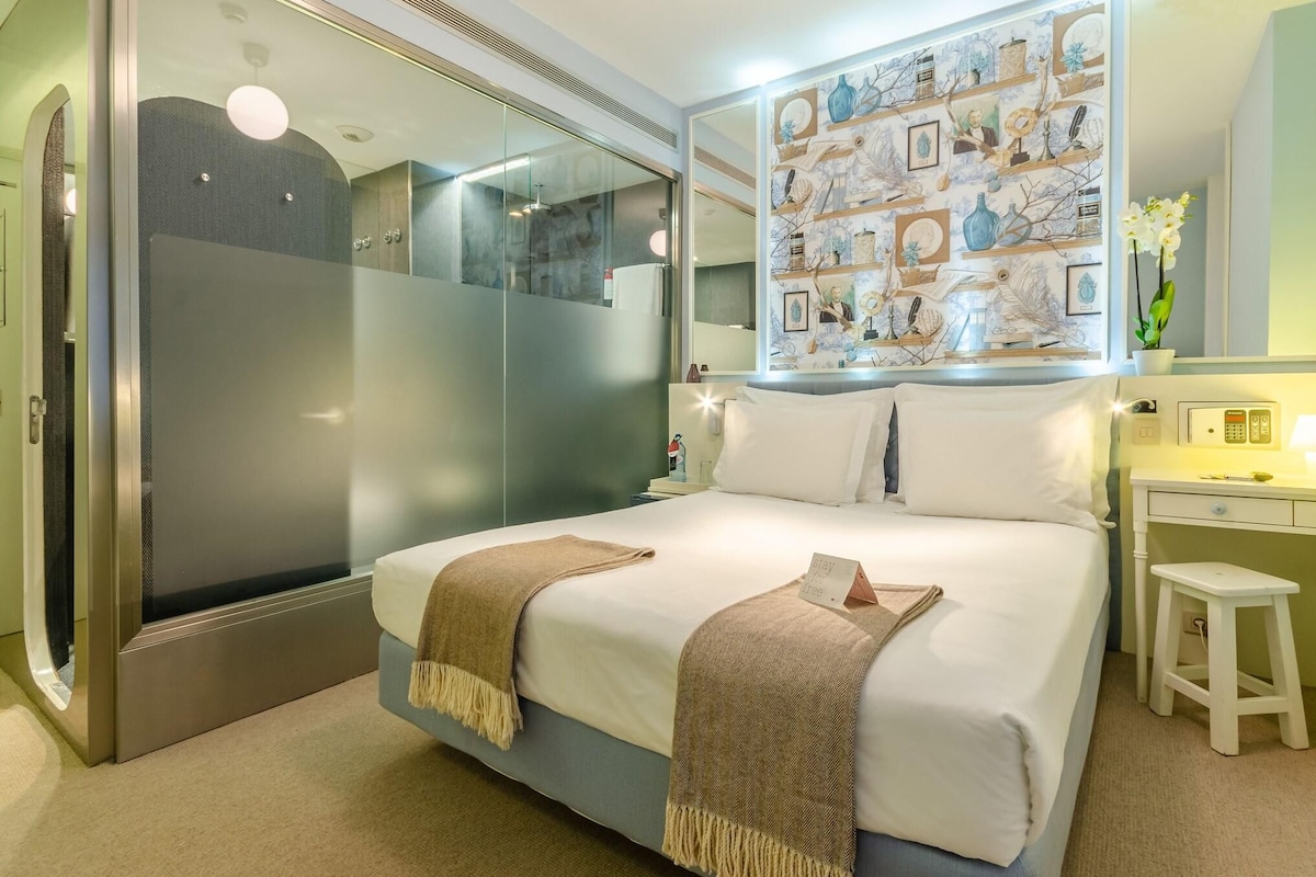 Une chambre d’hôtel moderne dotée d’un grand lit, d’une douche à parois de verre, d’une coiffeuse avec un miroir et d’un mur décoré d’œuvres d’art encadrées.