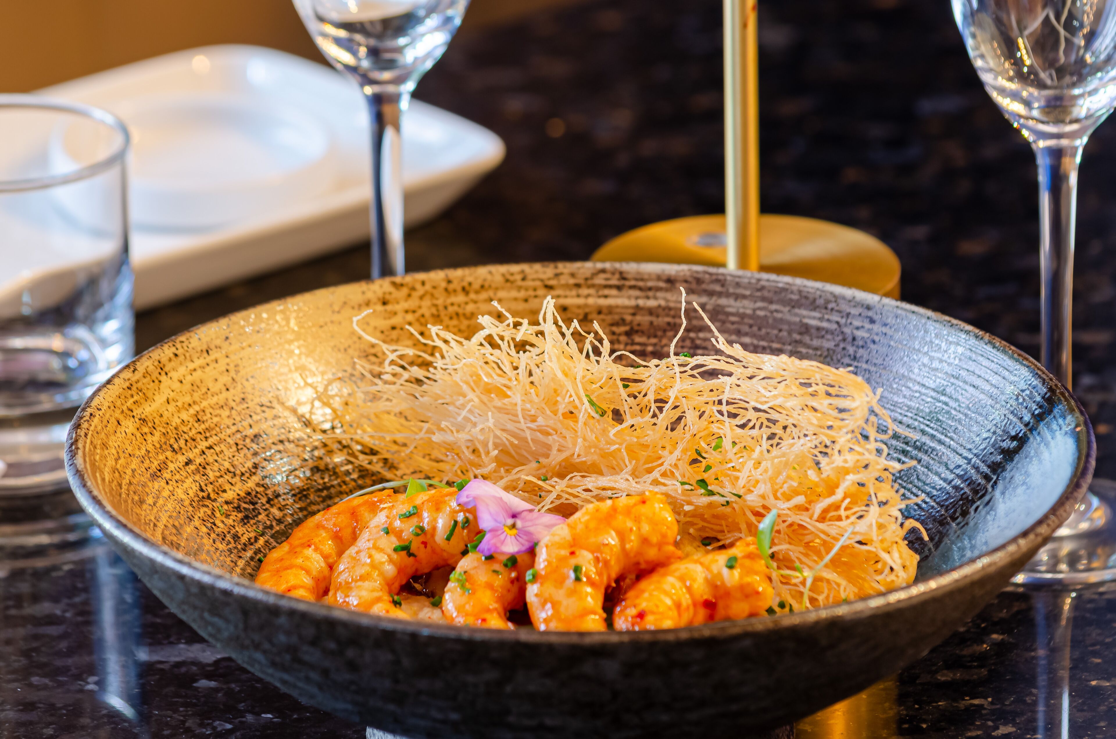 A bowl of food with noodles and shrimp, accompanied by a wine glass and a small plate.