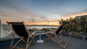 A rooftop terrace with two wooden lounge chairs and a small table, overlooking a body of water and a cityscape during sunset.