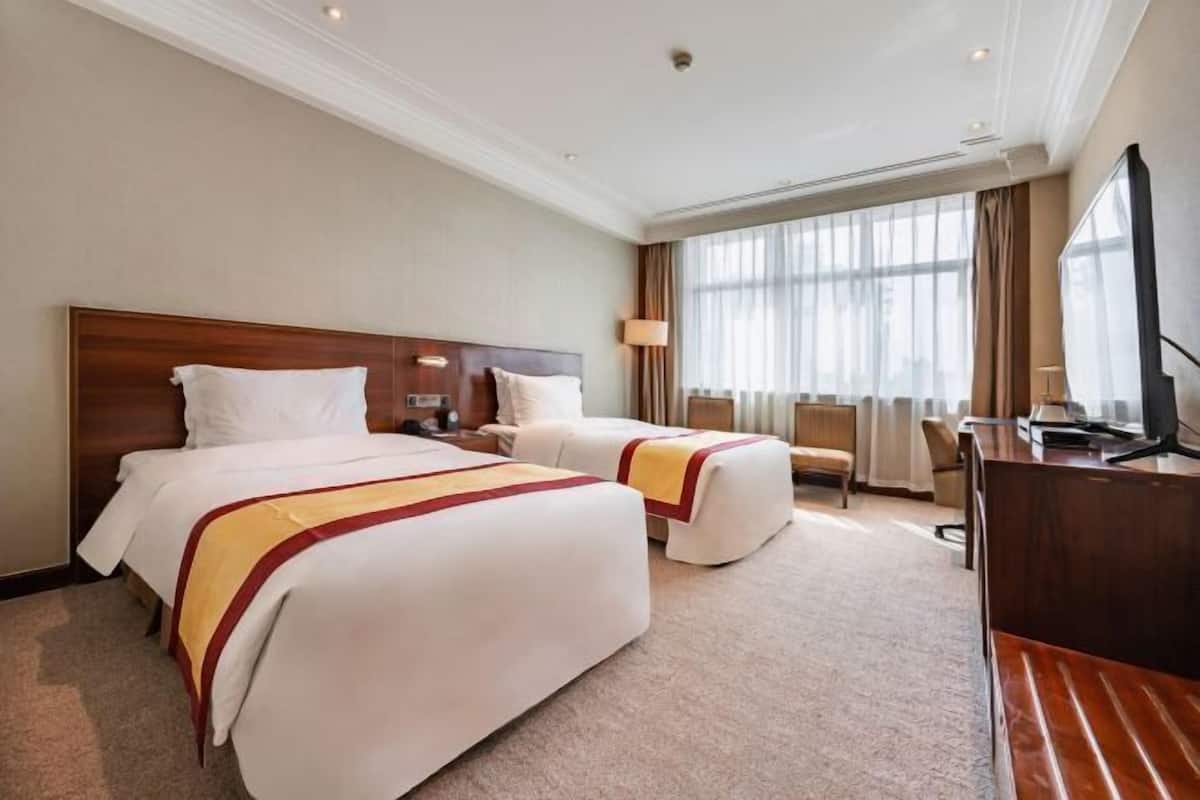 A hotel room with two beds, a desk, a chair, a TV, and a large window with curtains.