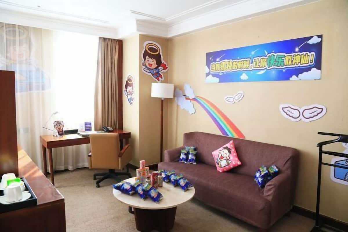 A hotel room with a brown sofa, a round coffee table with snacks, a desk with a lamp, and a wall with a cartoon character and a rainbow.