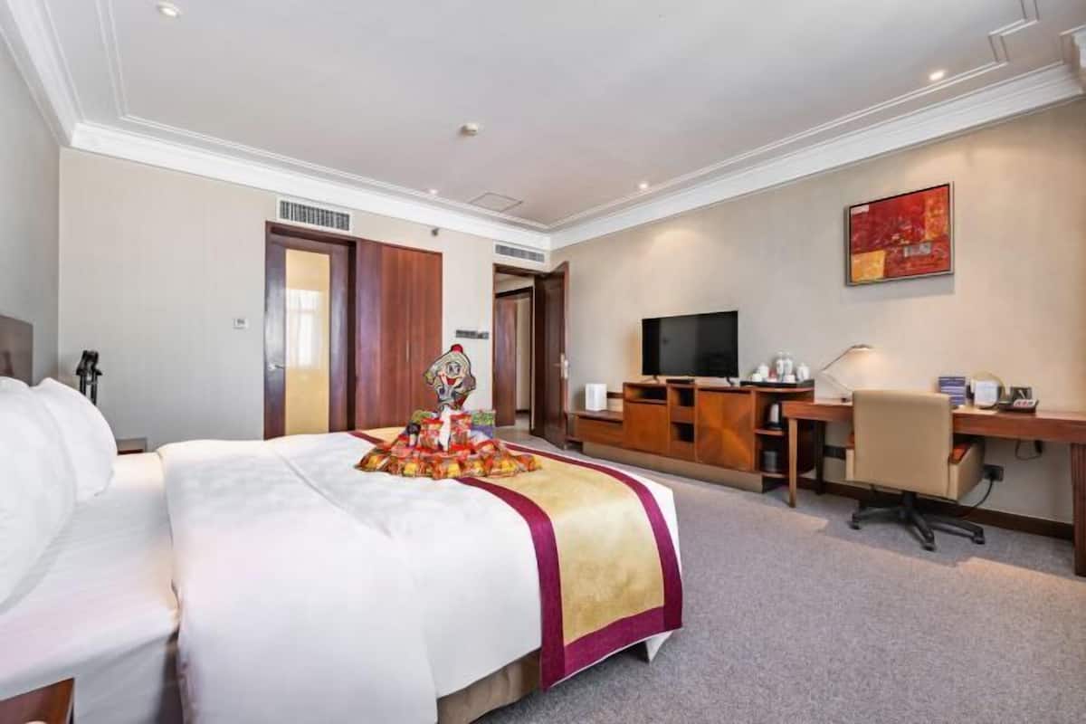 A hotel room with a bed, desk, chair, TV, and artwork on the wall.