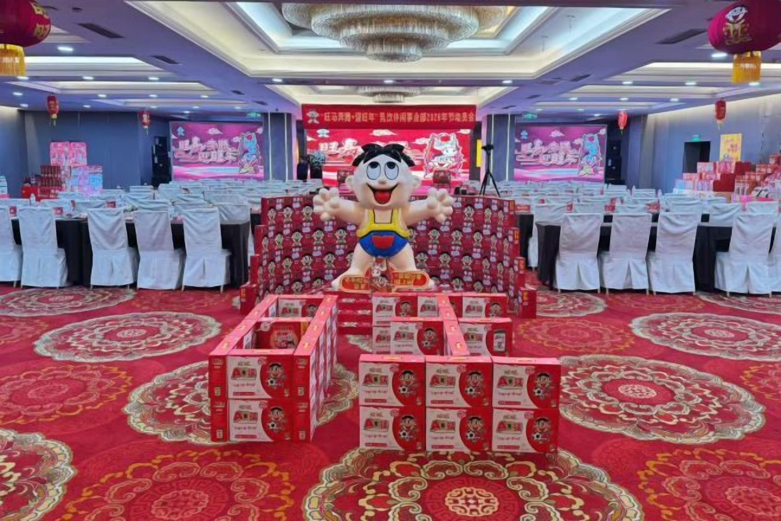 A banquet hall with round tables, a stage with a large mascot, and red boxes in the foreground.