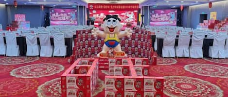 Un salón de banquetes con mesas redondas, un escenario con un gran personaje publicitario y cajas rojas en primer plano.