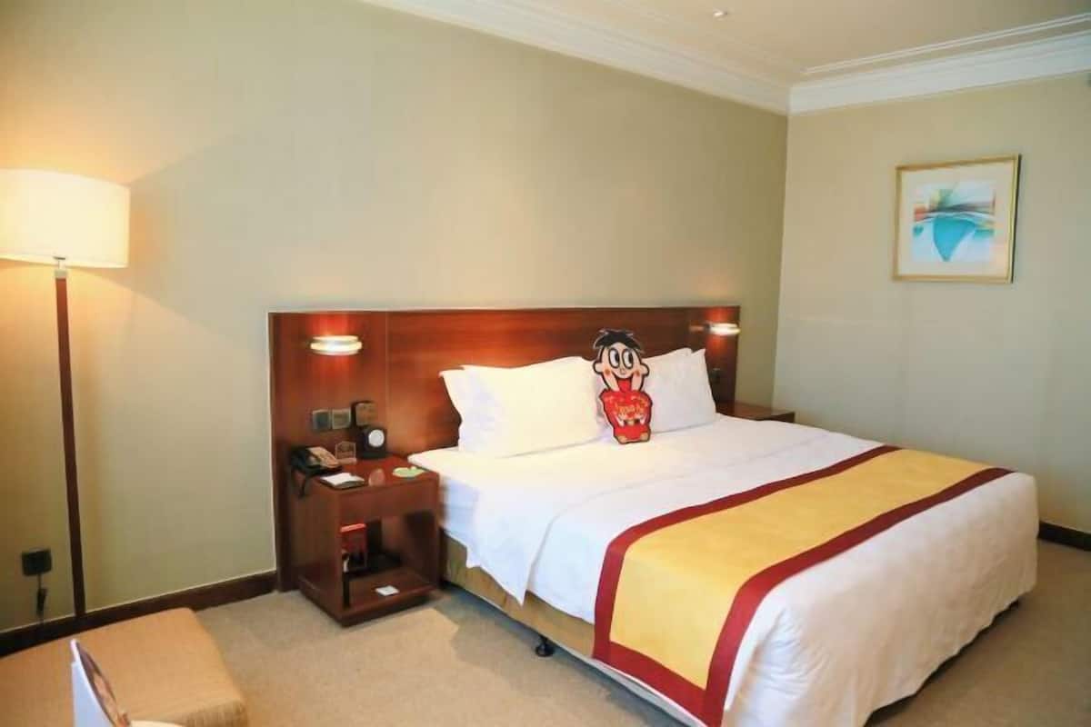 A hotel room with a large bed, a nightstand with a lamp, a small table, and a wall-mounted artwork.