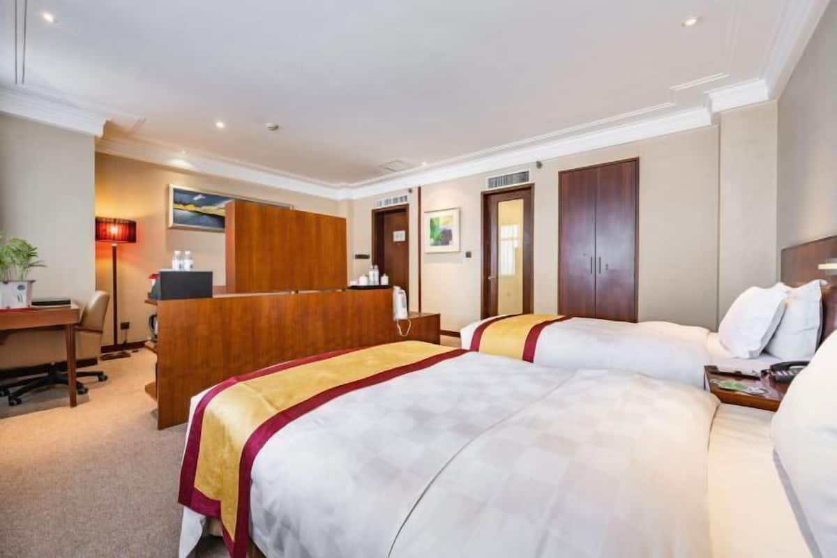A hotel room with a desk, two beds, a wooden reception desk, and a painting on the wall.