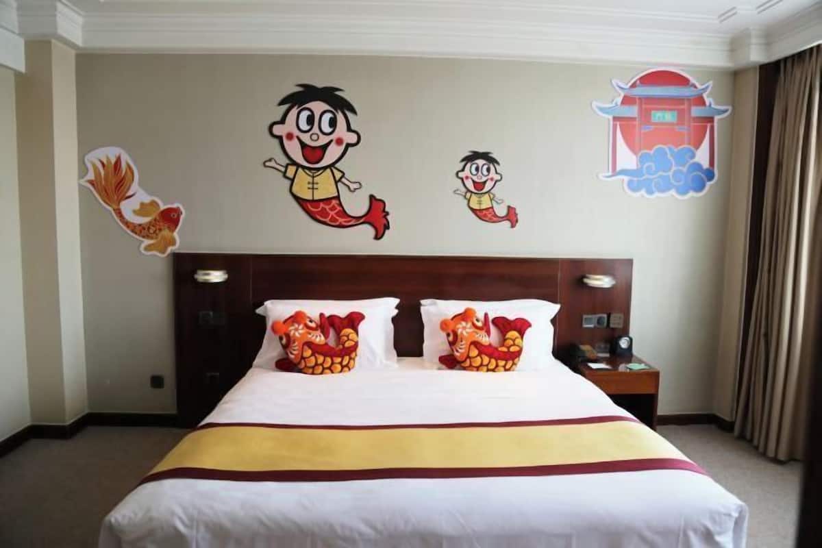 A hotel room with a large bed, two decorative pillows, a wooden headboard, a nightstand, and wall decorations including cartoon characters and a traditional building illustration.