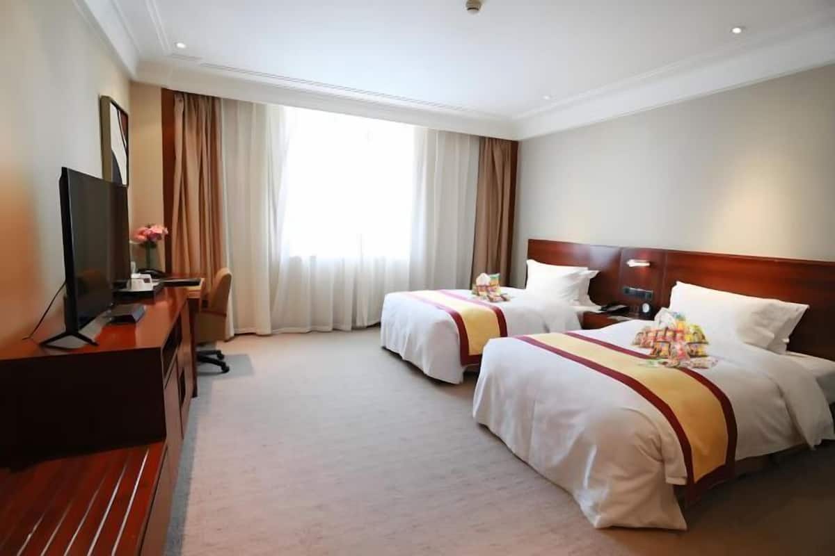 A hotel room with two beds, a TV, a desk, and a large window with curtains.