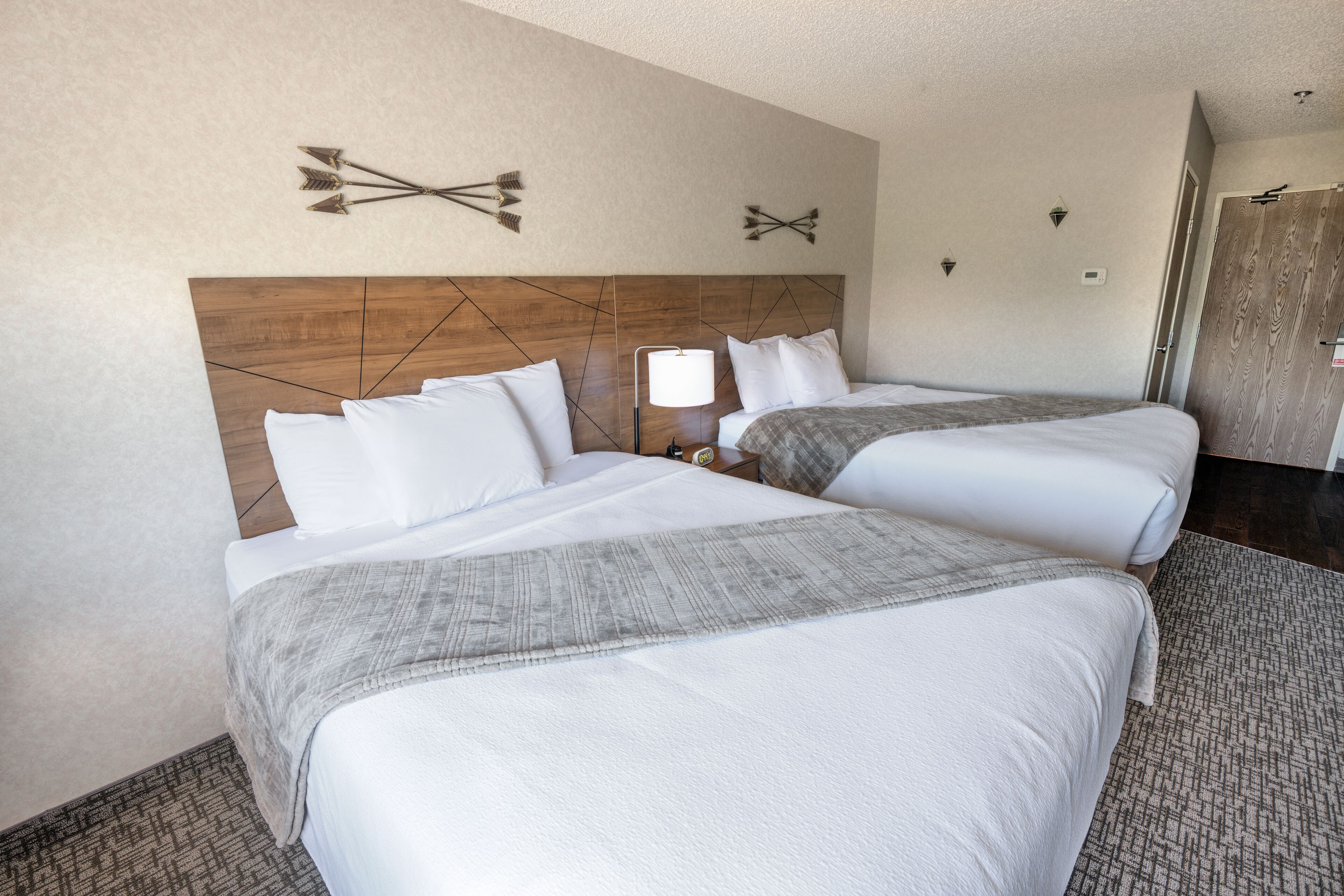 Habitación de hotel con dos camas, cabecero de madera, mesita de noche con lámpara y aplique de pared.