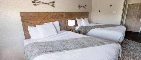 Habitación de hotel con dos camas, cabecero de madera, mesita de noche con lámpara y aplique de pared.
