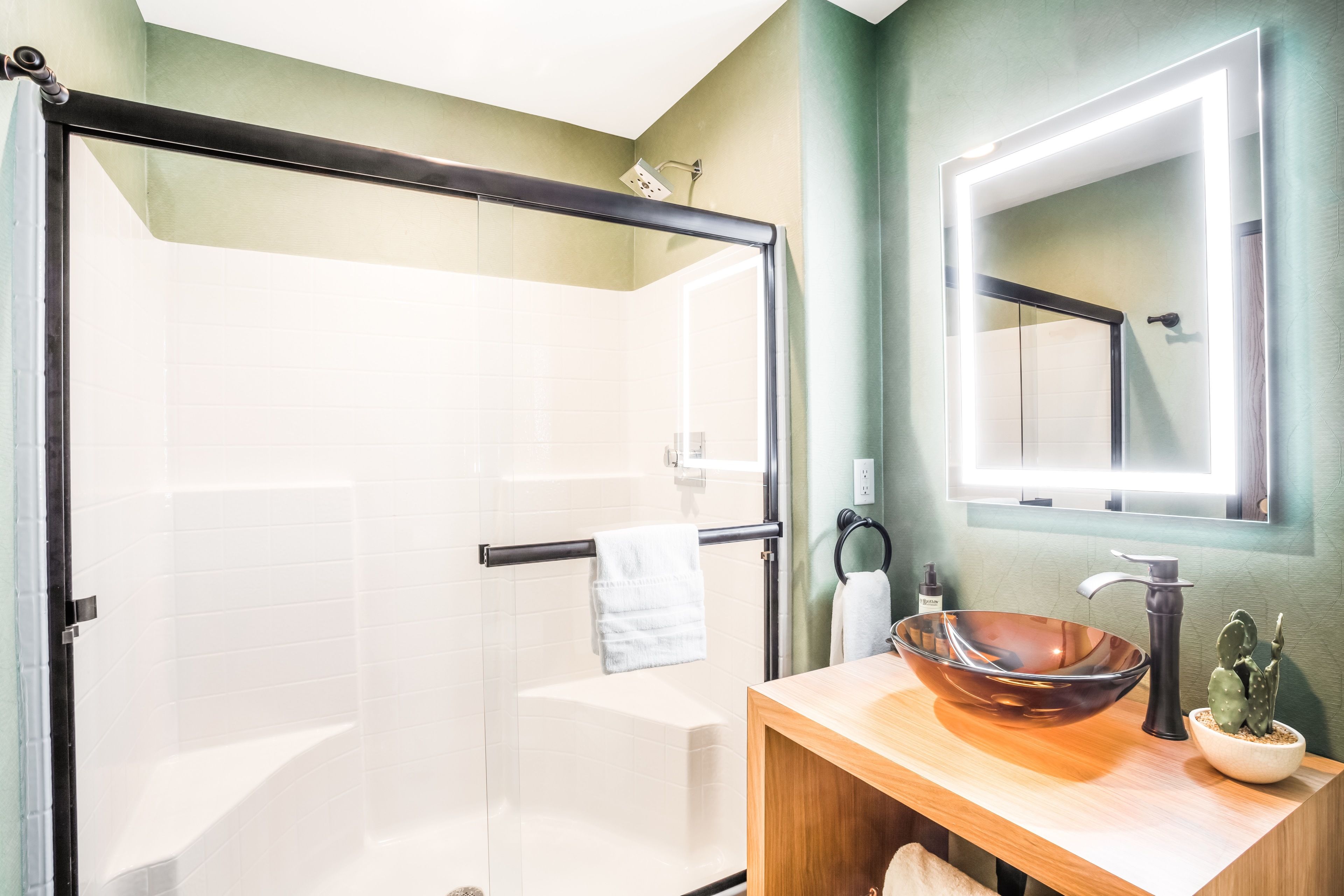 Un baño moderno con ducha envuelta en vidrio, tocador de madera con lavabo grande y espejo sobre el lavabo.