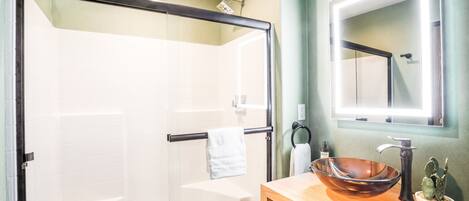 Un baño moderno con ducha envuelta en vidrio, tocador de madera con lavabo grande y espejo sobre el lavabo.