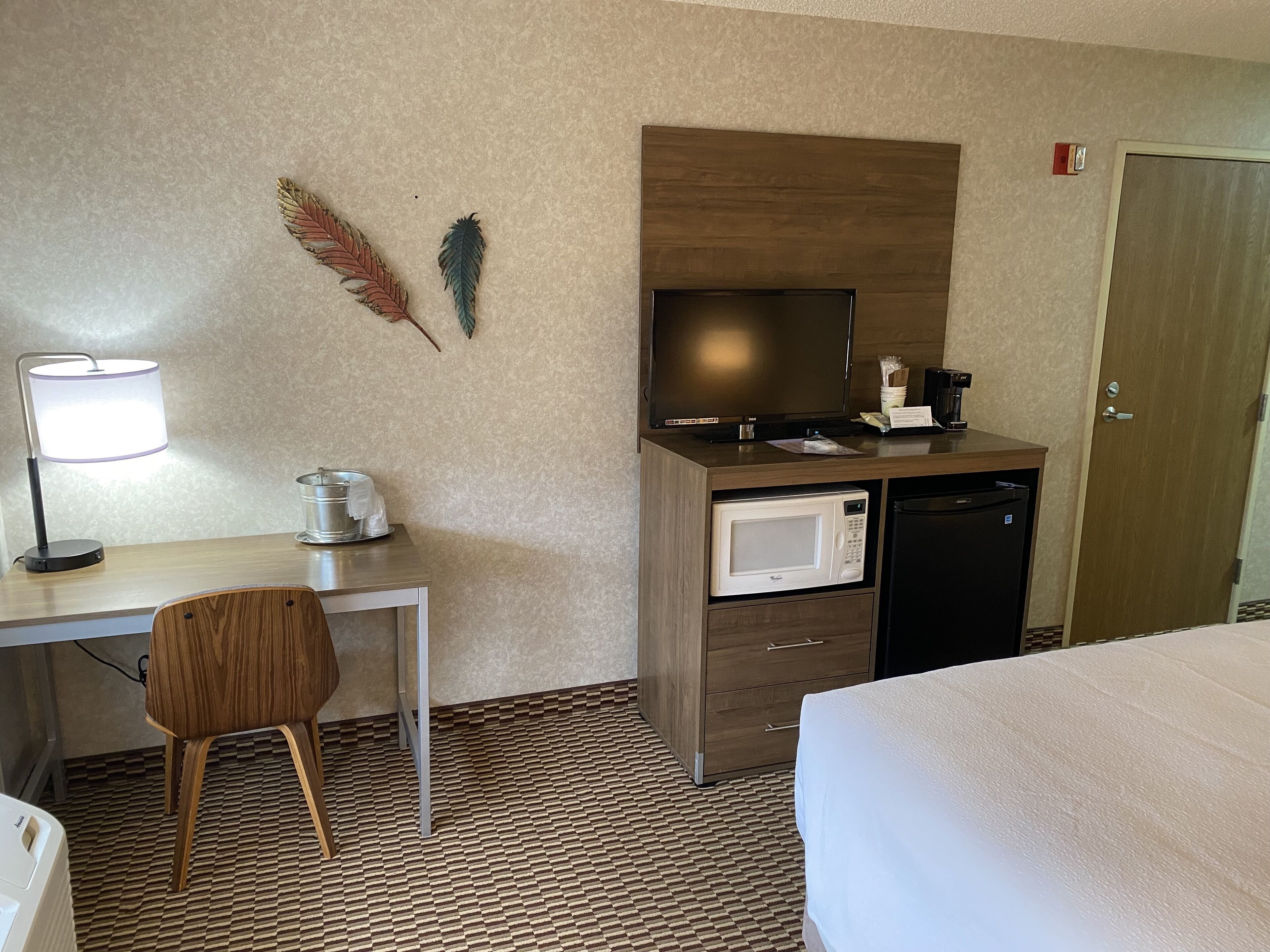 A hotel room with a bed, a desk with a lamp, a microwave, a television, and a door.