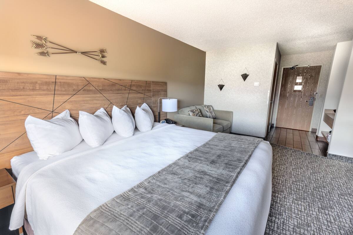 A hotel room with a large bed, a wooden headboard, a grey blanket, a bedside table, a lamp, a sofa, and a door.