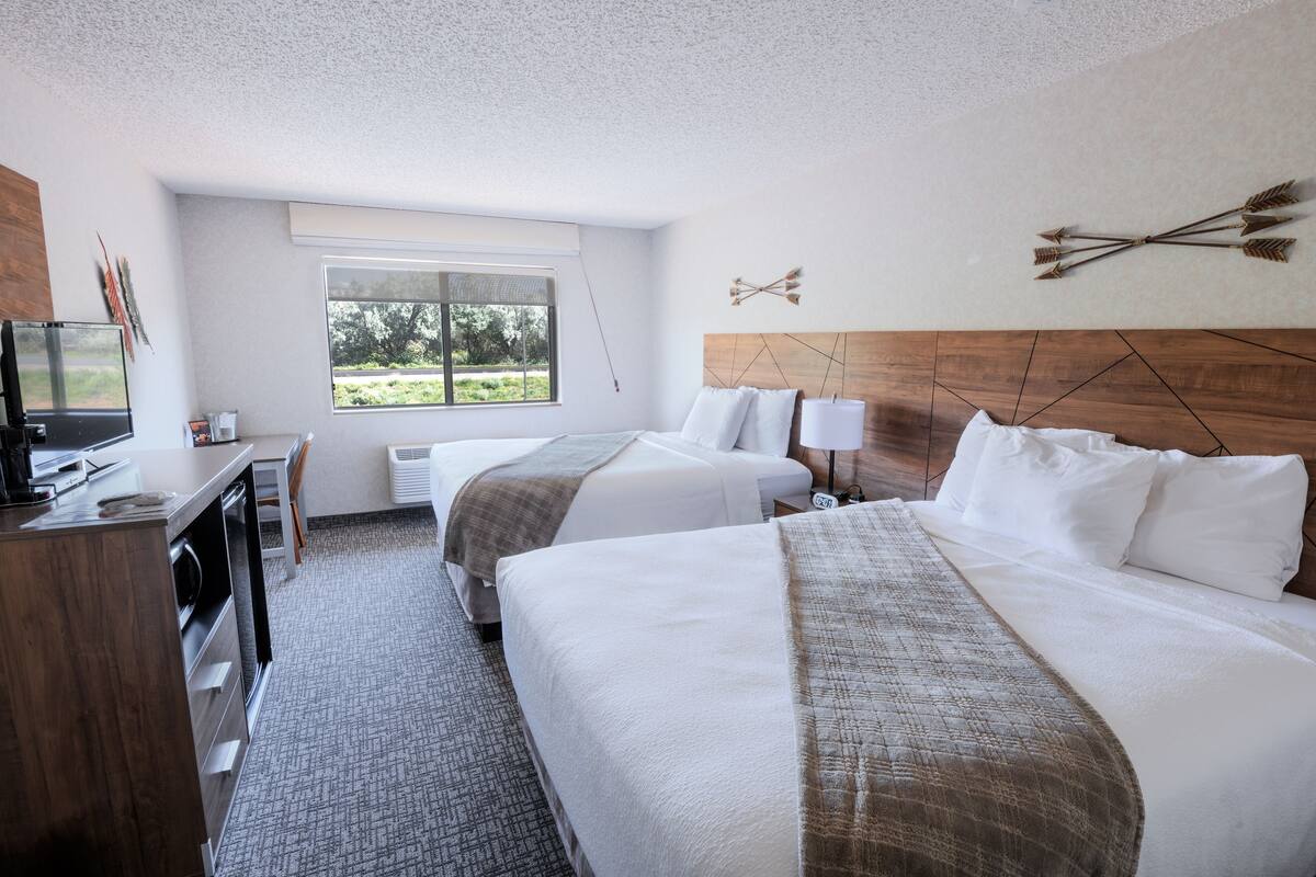 A hotel room with two beds, a desk, a television, and a window with a view of greenery.