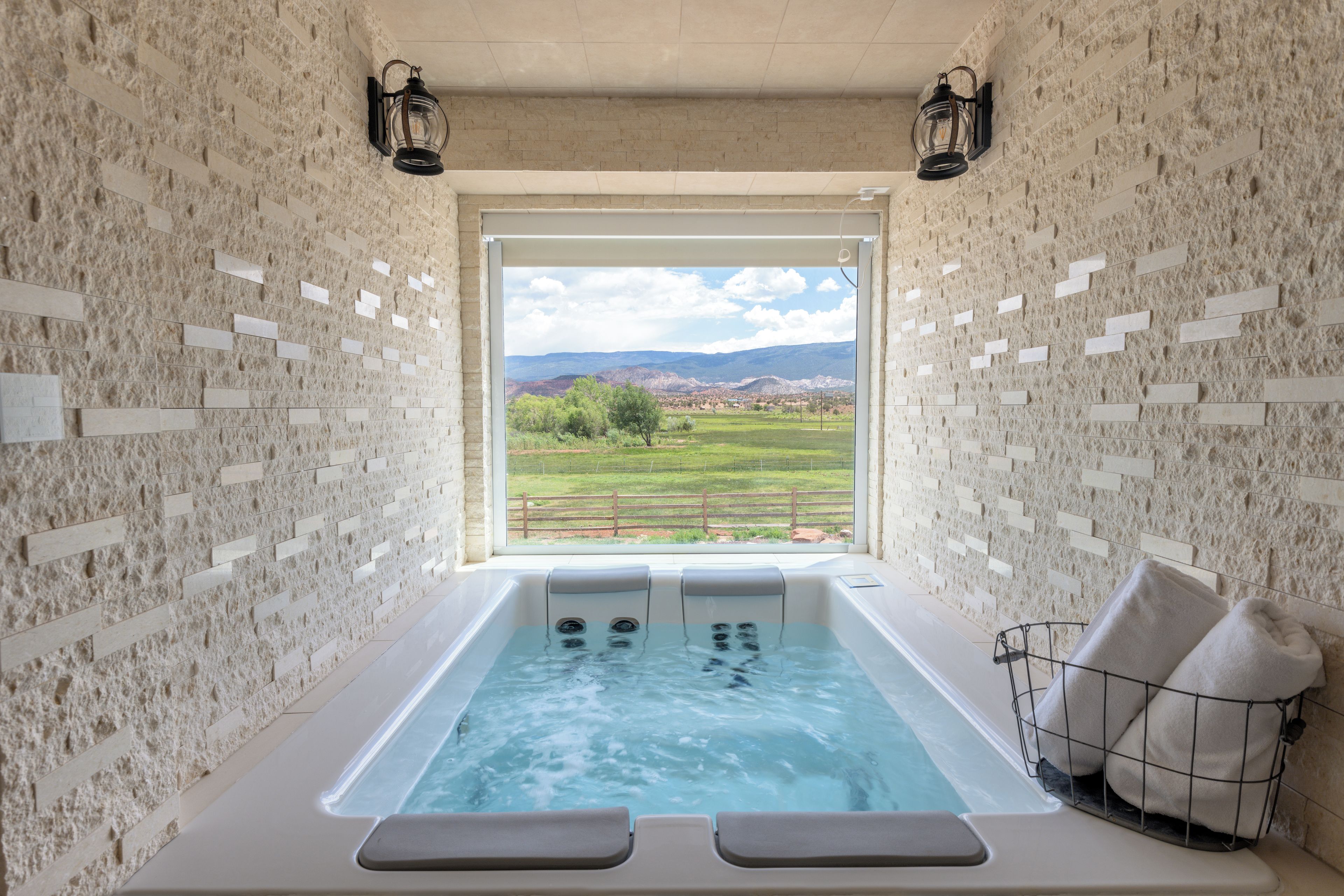 Une salle de spa avec un mur en pierre, une grande fenêtre, un jacuzzi et deux lampes fixées au mur.