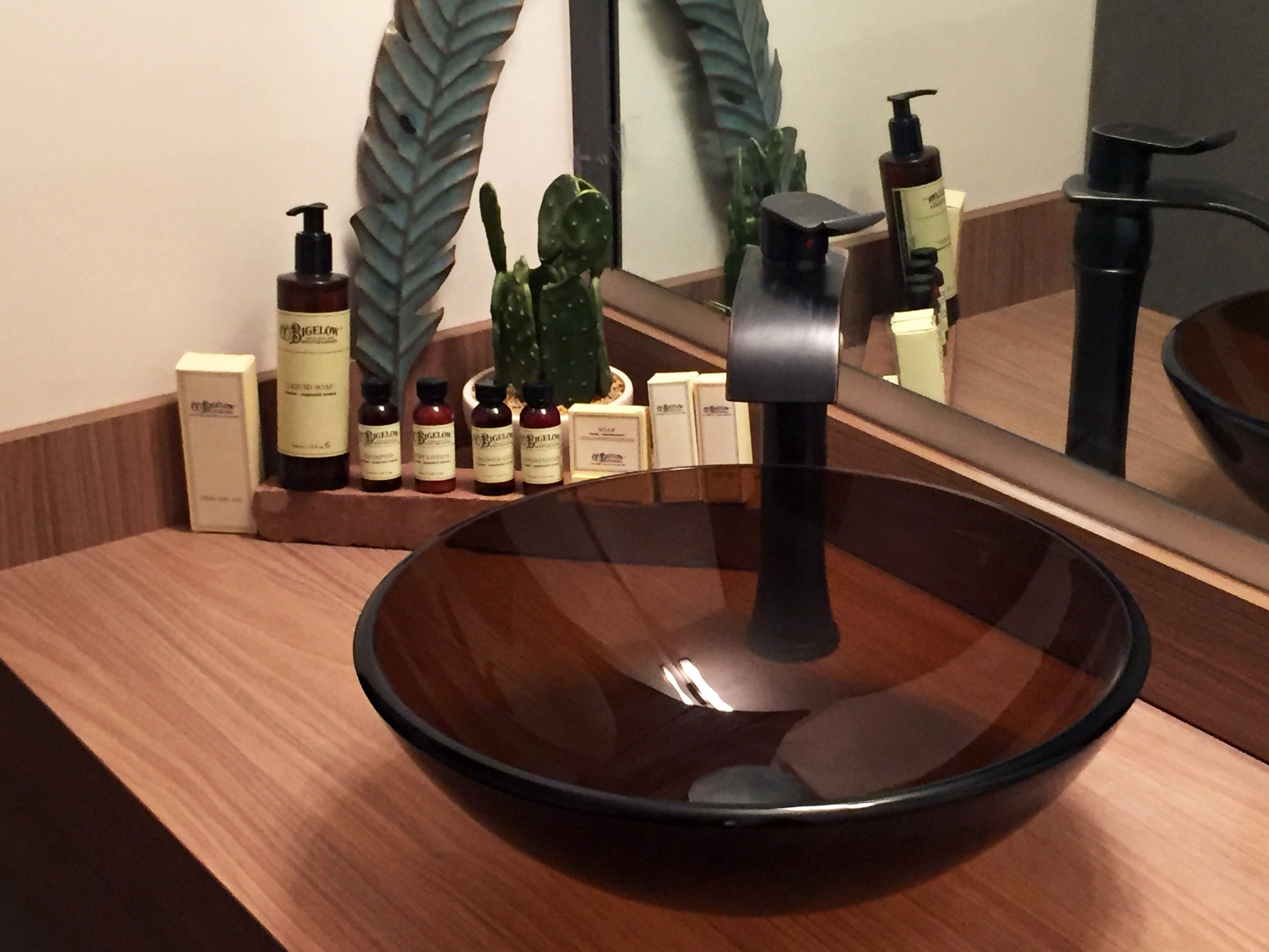 Un baño moderno con un lavamanos grande y de color oscuro, un espejo y distintos artículos de aseo sobre un estante de madera.