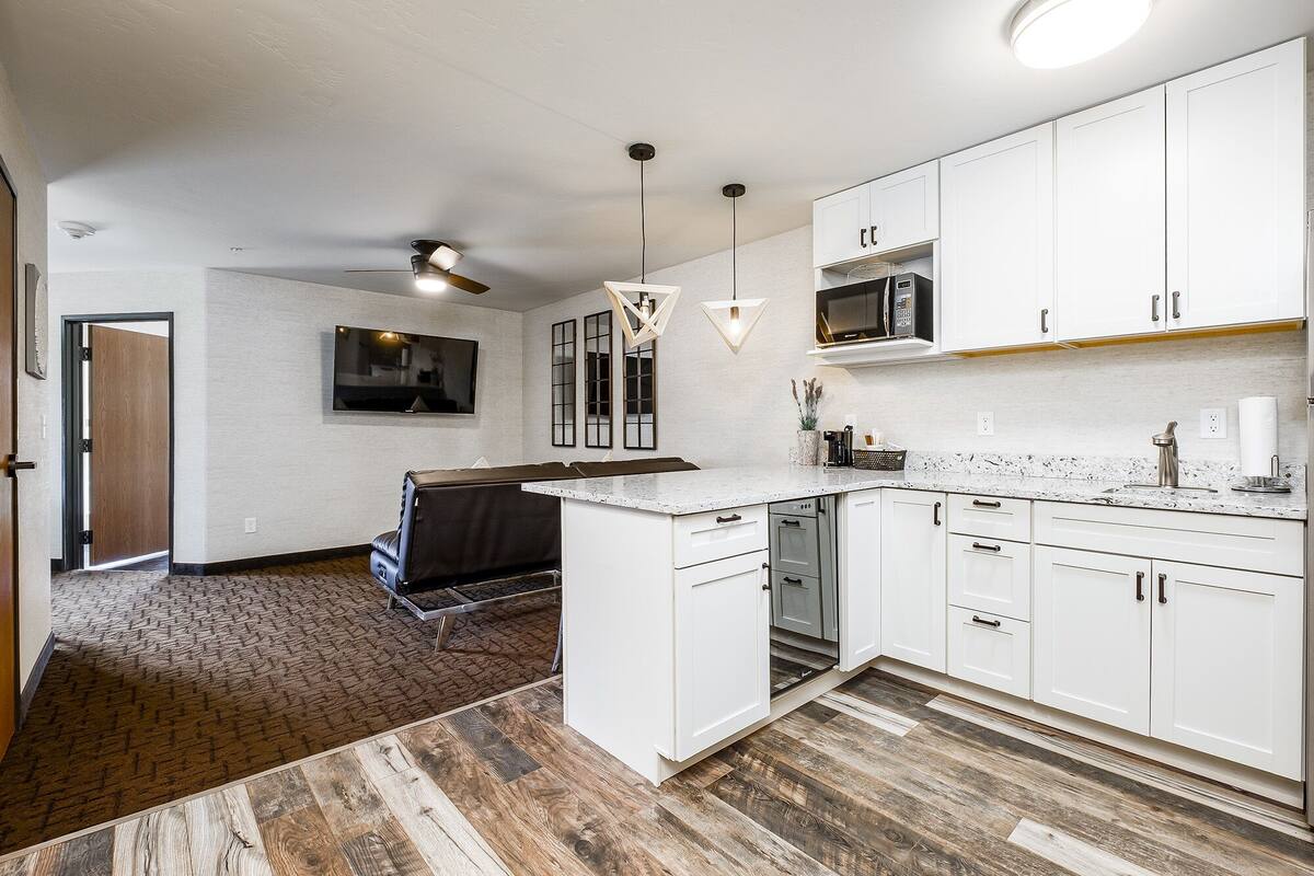 A modern kitchen with white cabinets, a built-in oven, and a microwave. There is a dining area with a brown leather couch and a TV mounted on the wall.