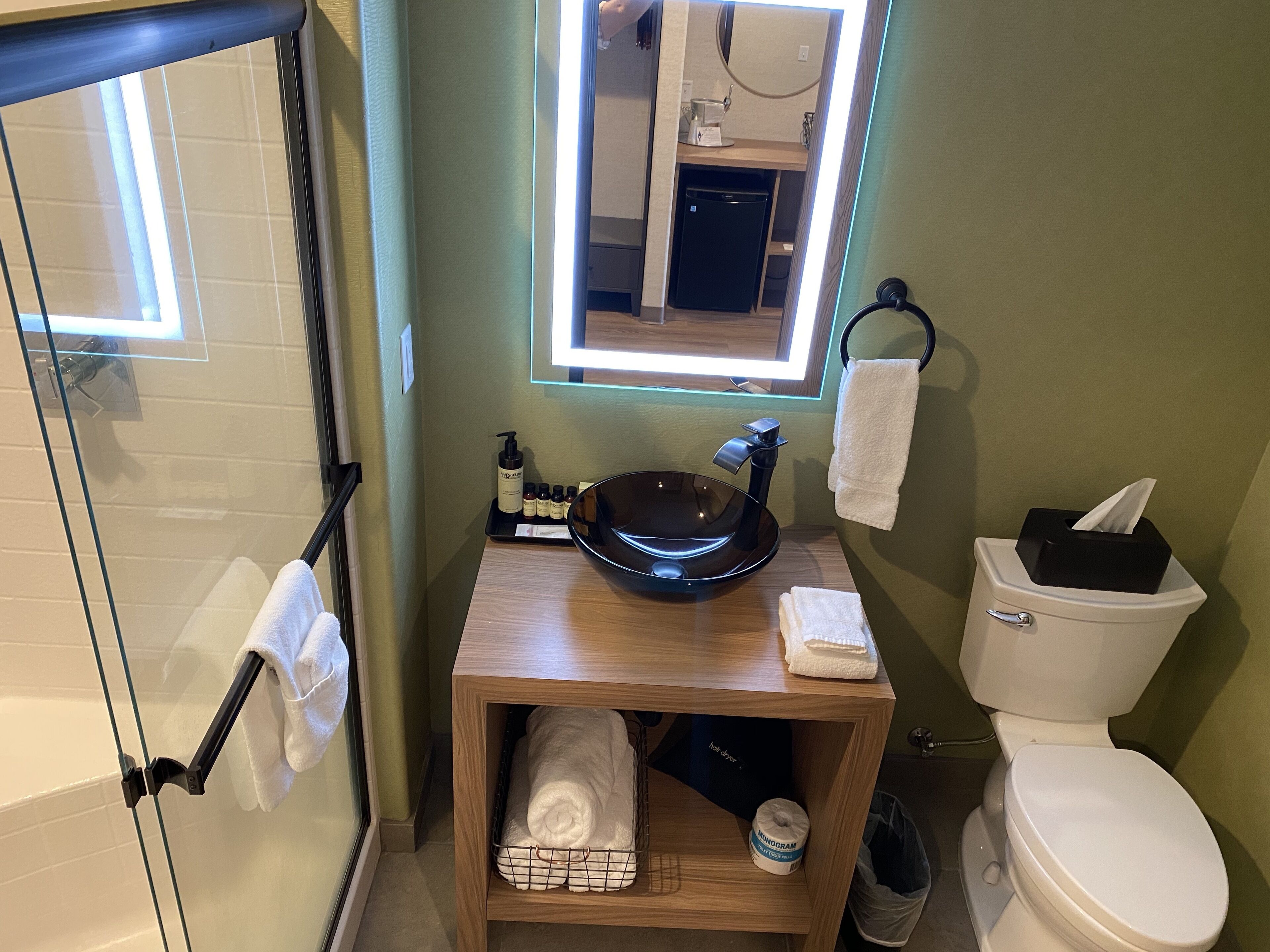 A bathroom with a glass shower enclosure, a wooden vanity with a sink, a toilet, and a small shelf with towels and toiletries.