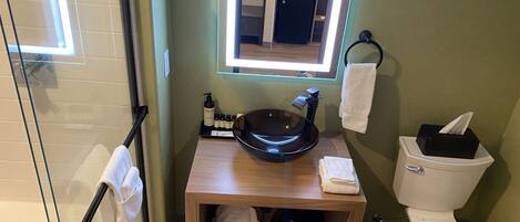 Een badkamer met een glazen douchecabine, een houten wastafelmeubel met een wastafel, een toilet en een klein plankje met handdoeken en toiletartikelen.
