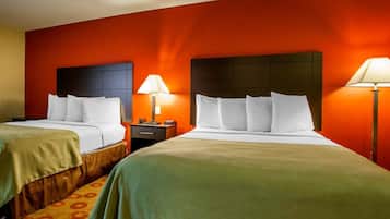 A hotel room with two beds, each with white bedding and a green throw blanket. There are two bedside tables with lamps, and the walls are painted orange.