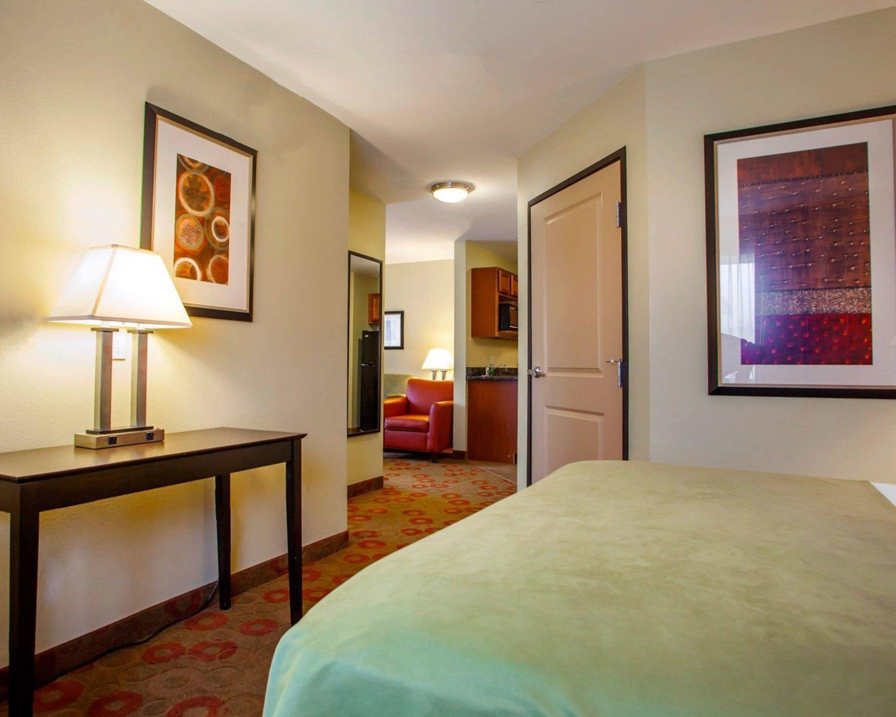 A hotel room with a bed, a desk with a lamp, a red chair, and a kitchen area visible through an open door.
