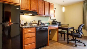 Una cocina compacta con armarios de madera, un refrigerador negro, lavavajillas, cafetera y microondas. Hay una mesa de comedor con sillas y una lámpara colgante arriba.