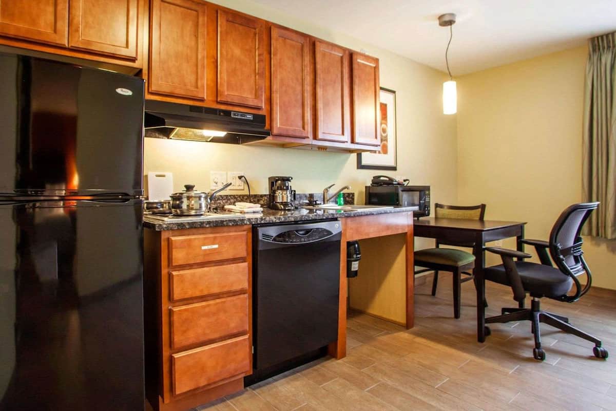 A compact kitchen with wooden cabinets, a black refrigerator, a dishwasher, a coffee maker, and a microwave. There's a dining table with chairs and a pendant light above it.