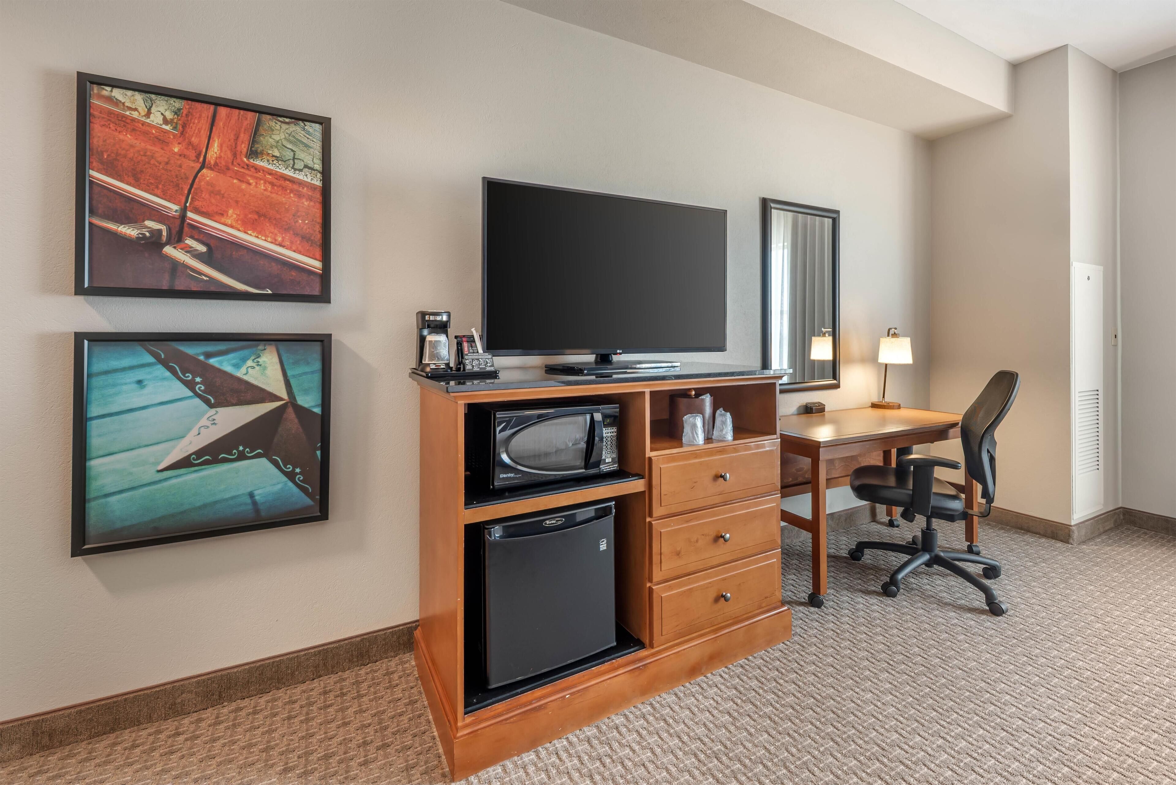 Une chambre d’hôtel équipée d’un téléviseur à écran plat, d’un petit réfrigérateur, d’un four à micro-ondes, d’un bureau avec une chaise et de deux œuvres d’art encadrées accrochées au mur.