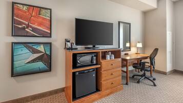 A hotel room with a flat-screen TV, a small refrigerator, a microwave, a desk with a chair, and two framed artworks on the wall.