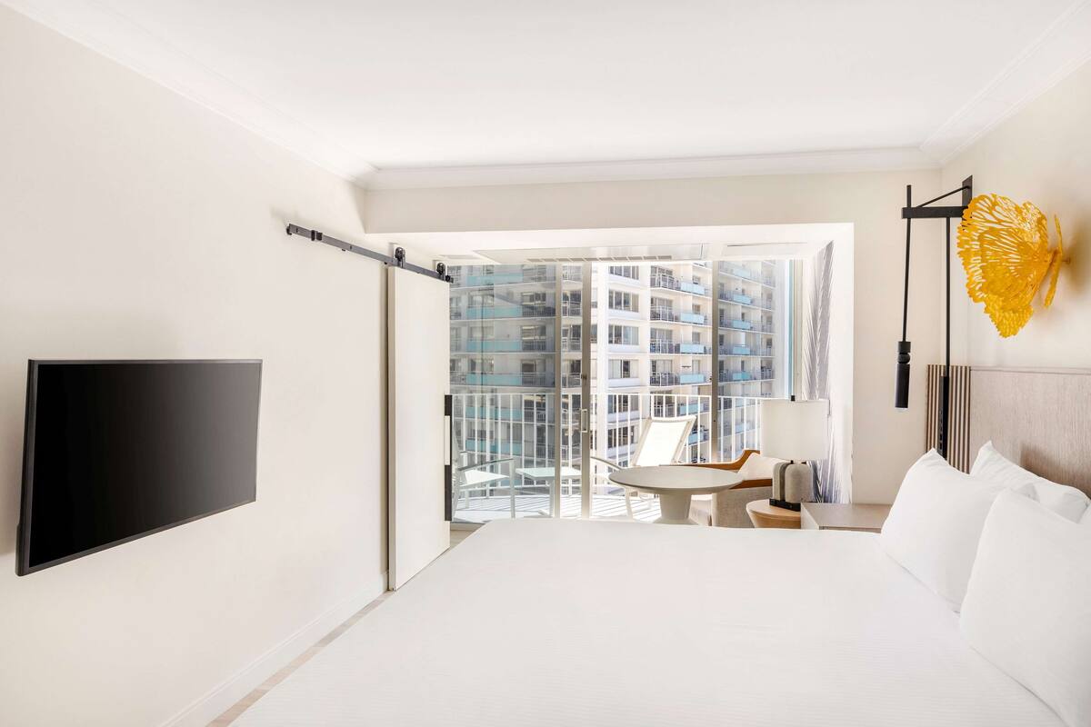 A modern hotel room with a large bed, a flat-screen TV, a balcony with a view of high-rise buildings, and a unique yellow wall decoration.