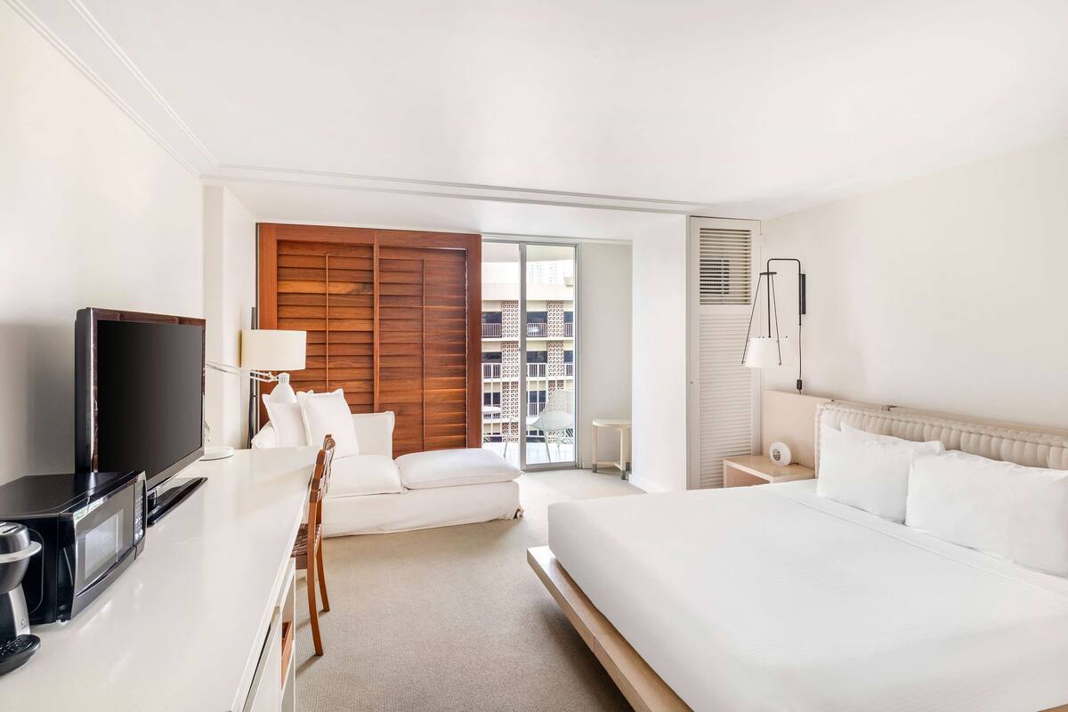 A hotel room with a bed, a sofa, a TV, a lamp, and a view of the city.