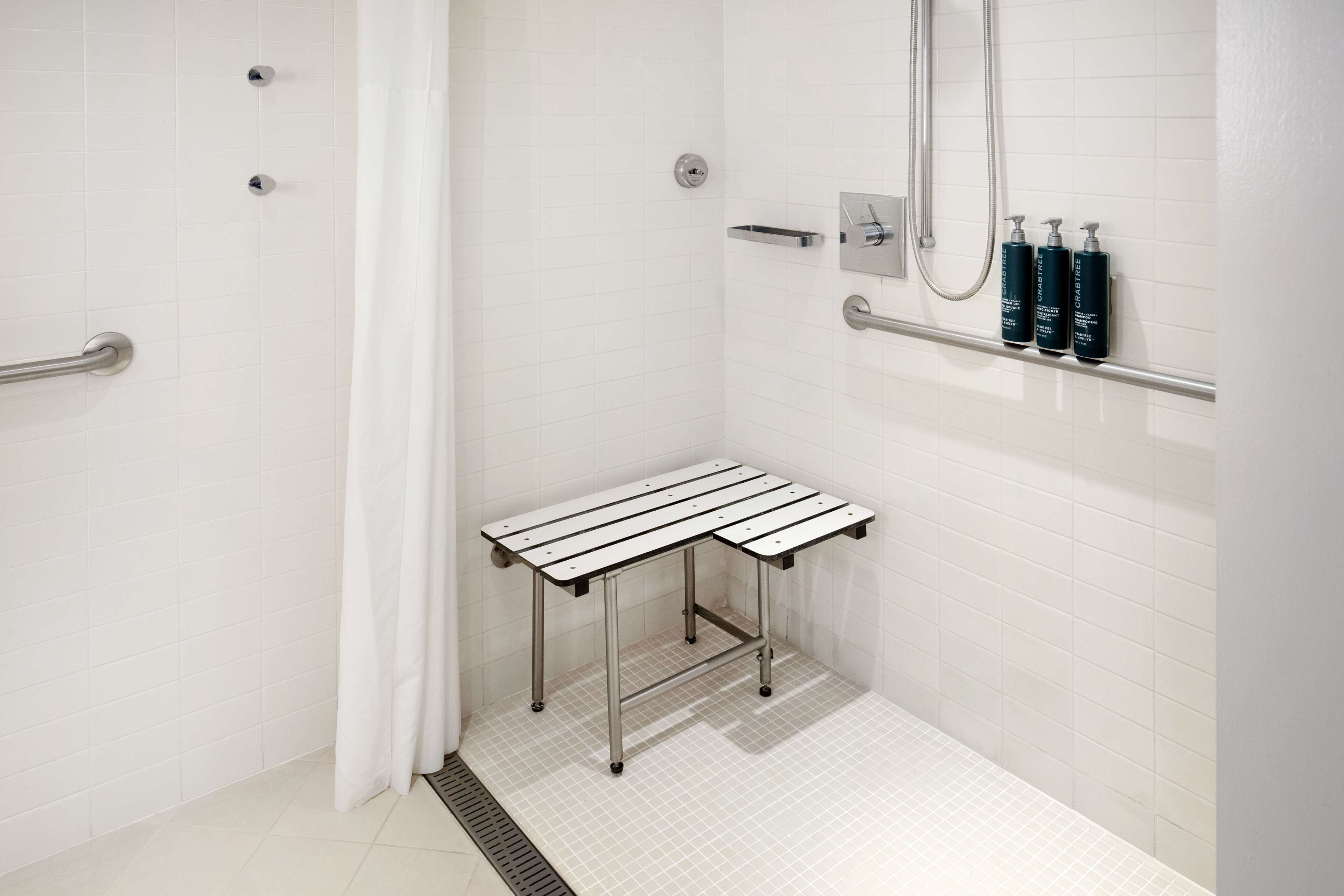 Een moderne badkamer met witte tegels, een douche met handdouche, een handgreep en een klein tafeltje met een zwart-wit gestreept blad.