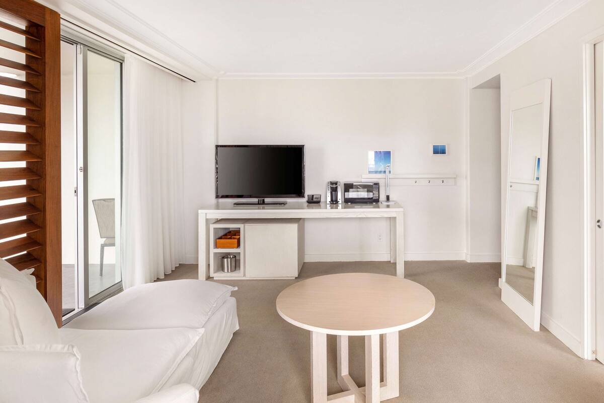 A modern hotel room with a white sofa, a round table, a flat-screen TV, a minibar, and a sliding glass door.