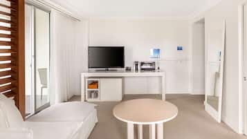 A modern hotel room with a white sofa, a round table, a flat-screen TV, a minibar, and a sliding glass door.