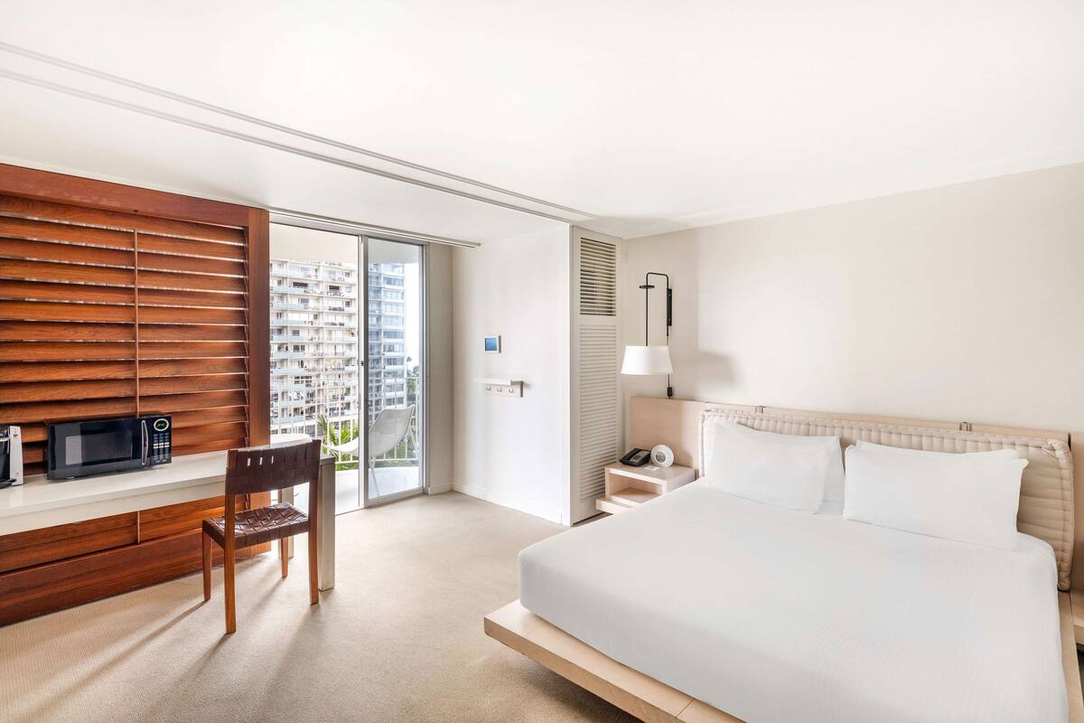 A hotel room with a bed, a desk with a chair, a microwave, a lamp, and a view of a cityscape through a sliding door.
