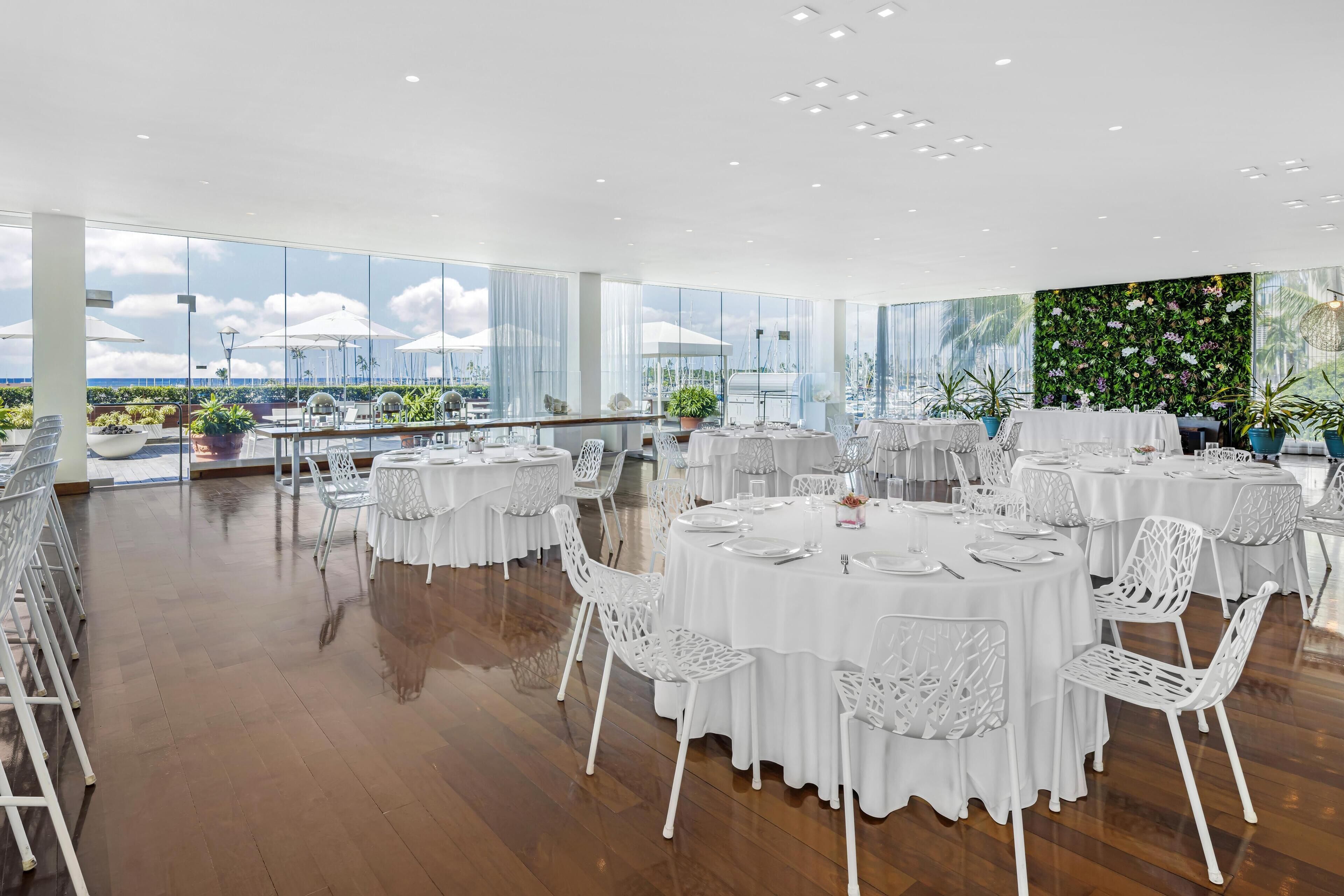 Un'ampia sala da pranzo interna con tavoli rotondi e sedie bianche, grandi finestre che offrono vista sull'esterno e una parete verde con piante.