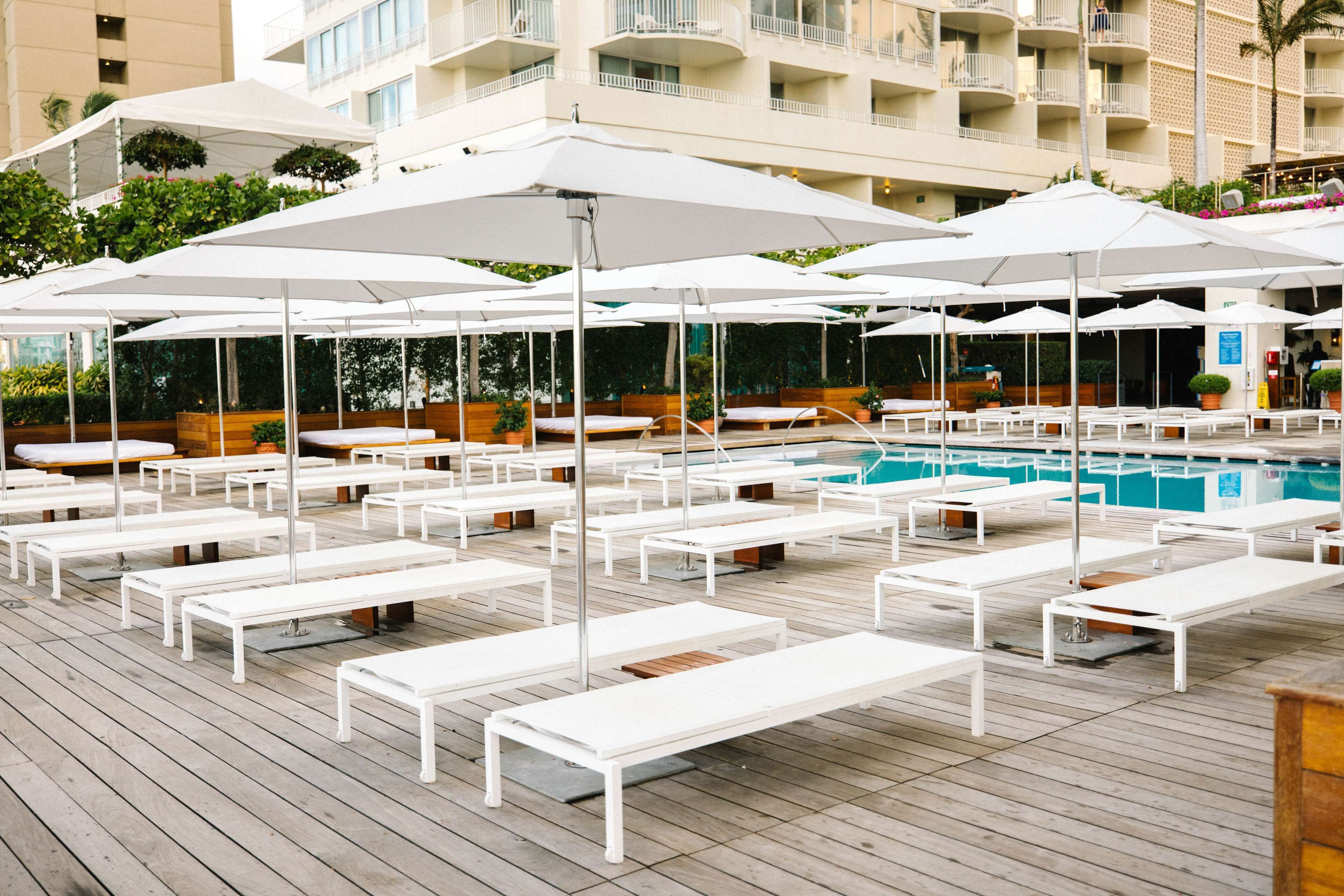 A poolside area with white lounge chairs and umbrellas, a pool, and a building with balconies in the background.