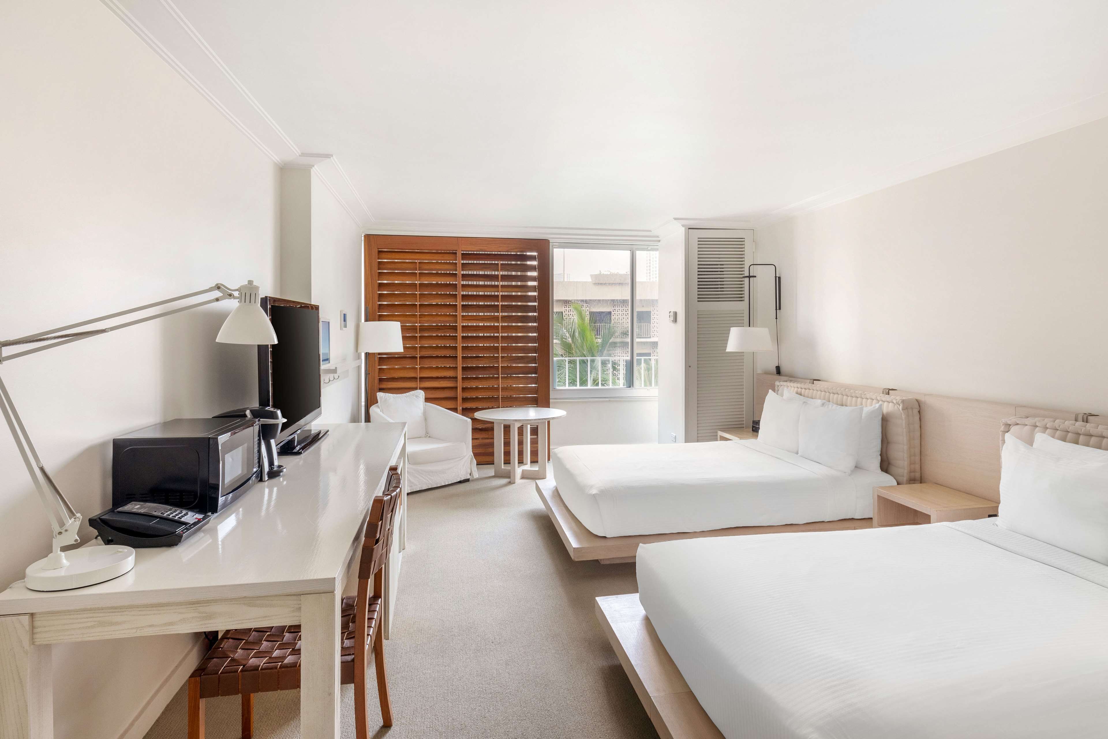 A hotel room with two beds, a desk with a computer, a TV, and a window with blinds.