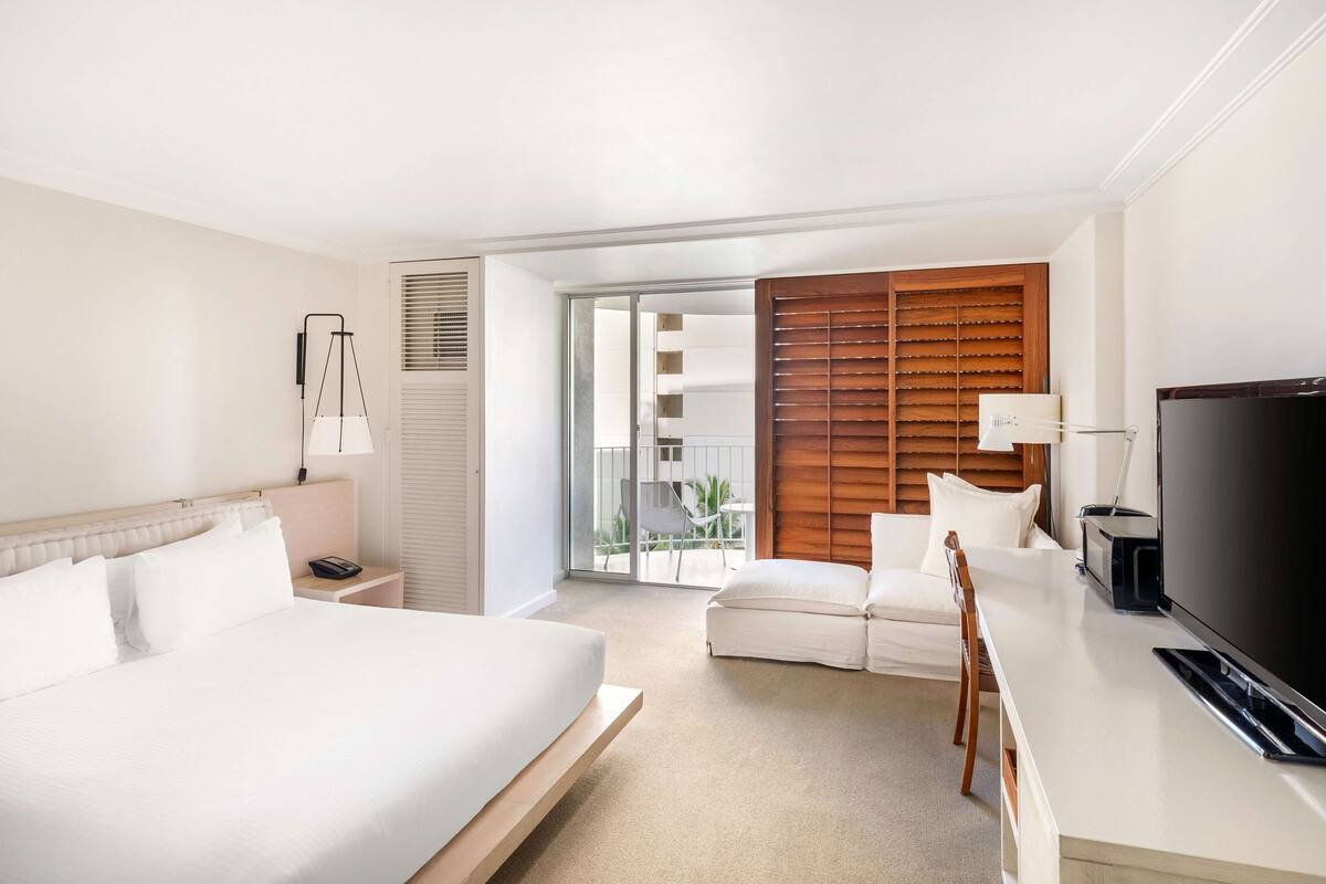 A hotel room with a bed, a desk with a TV, a chair, and a sliding door to a balcony.