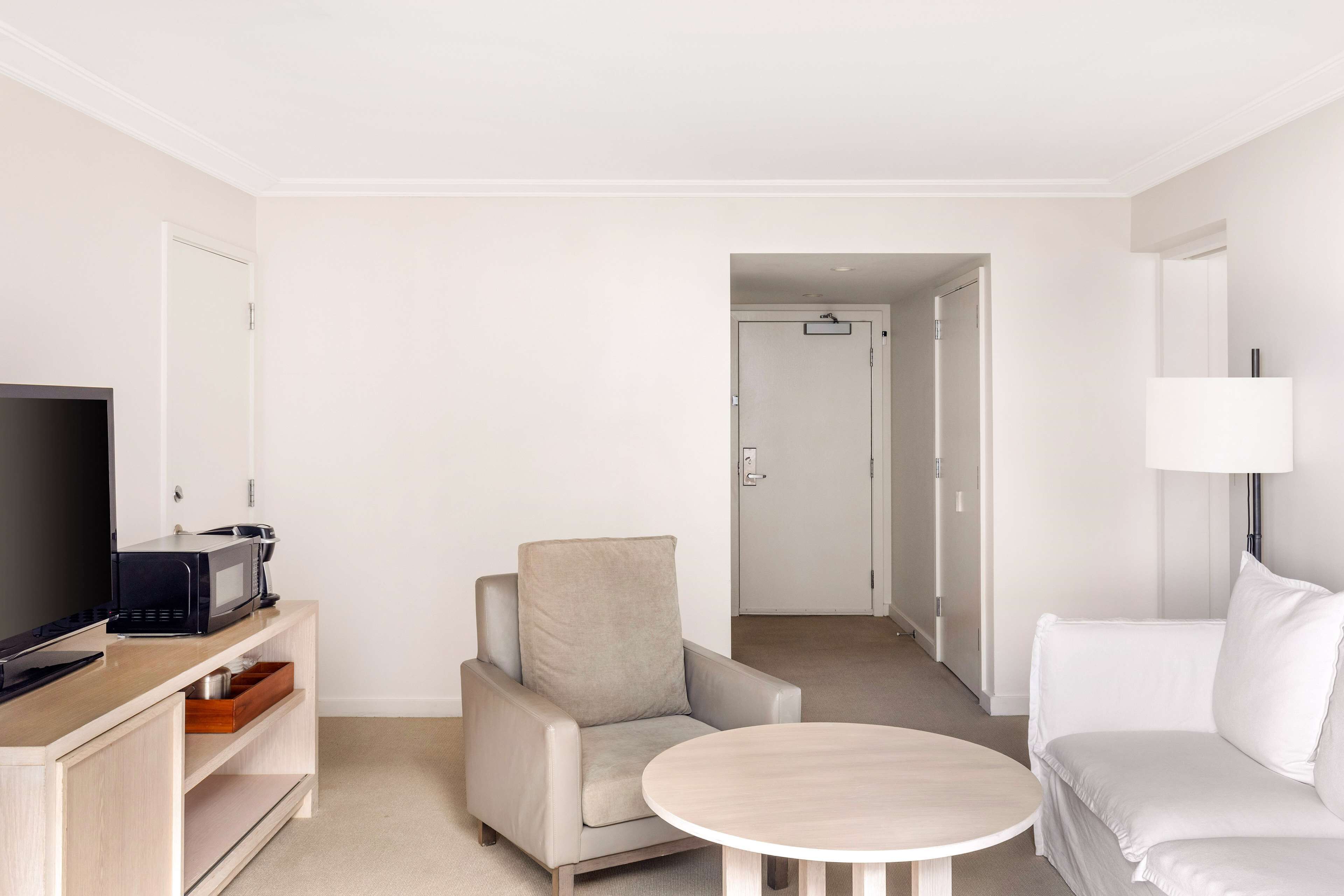 Habitación de hotel moderna con televisión de pantalla plana, un sillón color beige, un sofá blanco y una mesa redonda de madera.