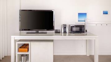 A white desk with a flat-screen TV, a coffee maker, and a microwave. There are storage drawers and a small trash can under the desk.