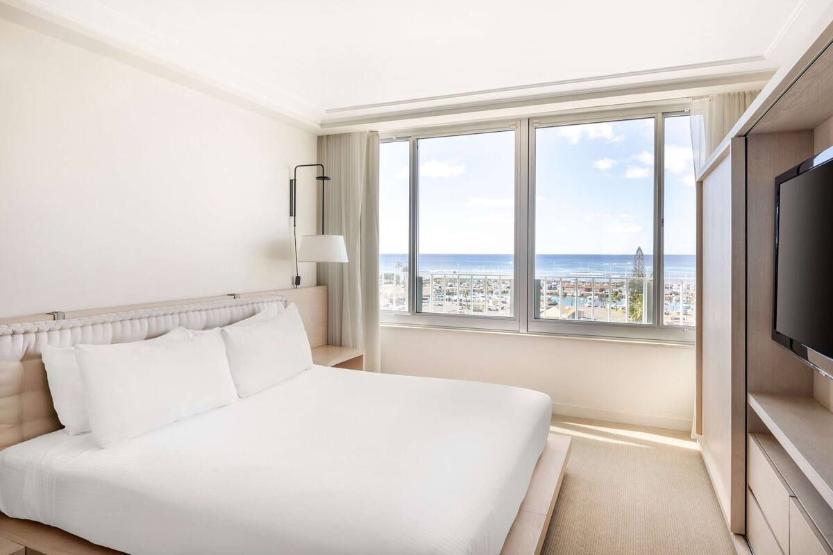 A hotel room with a large bed, a TV, and a view of the city and sea.