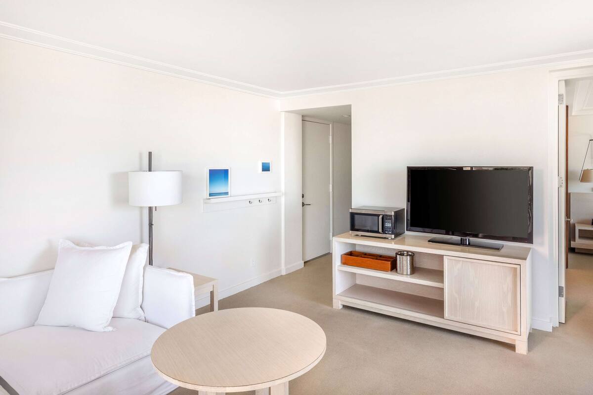 A hotel room with a white sofa, a round table, a TV on a wooden stand, a lamp, and a microwave.