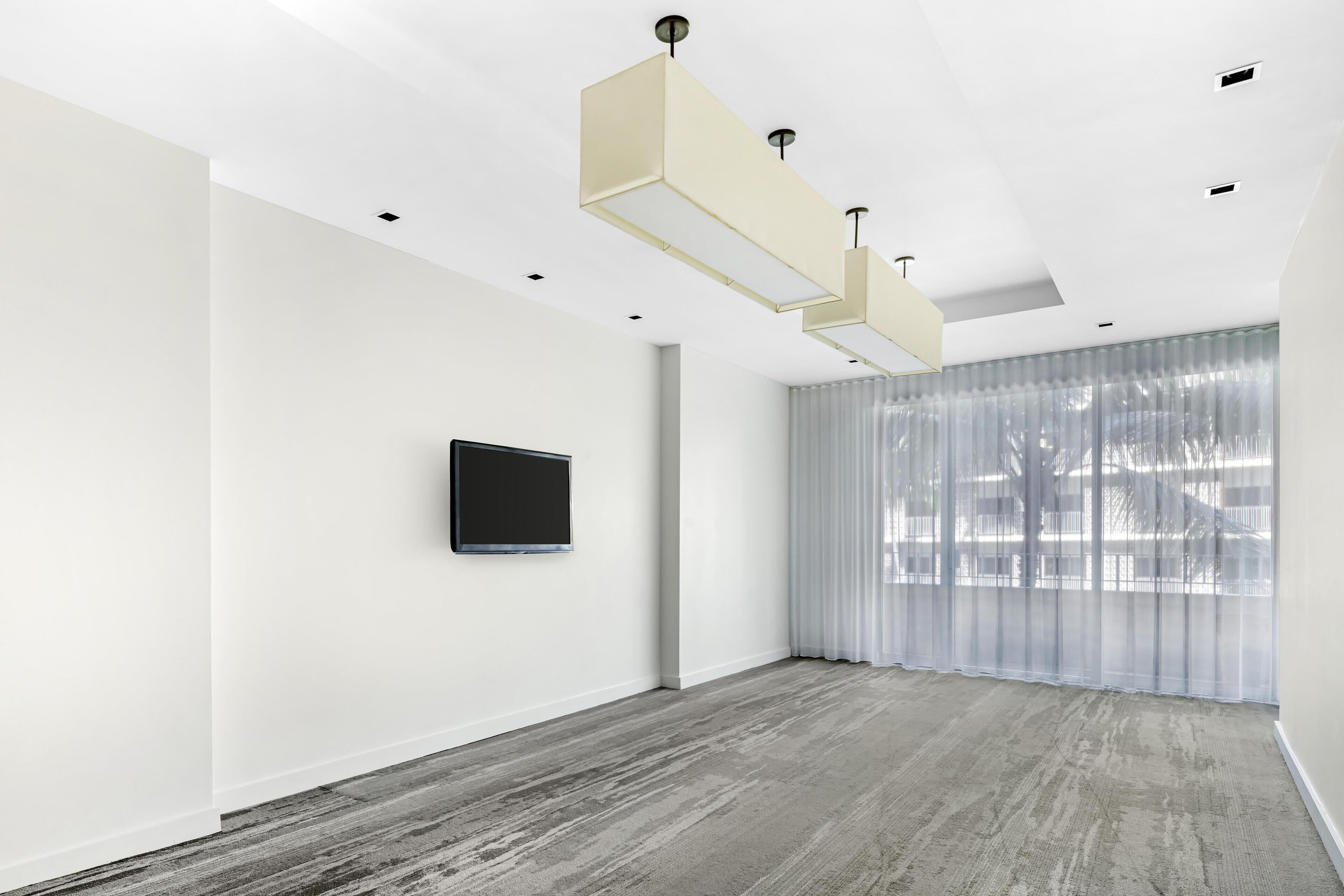 A spacious room with a large window, a flat-screen TV mounted on the wall, and a modern ceiling light fixture.