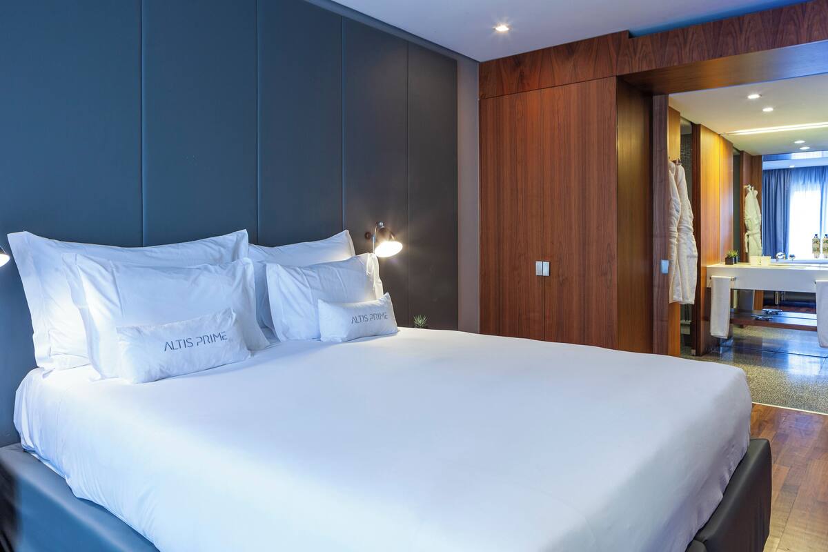 Ein großes Bett mit weißer Bettwäsche und Kissen mit dem Aufdruck "ALTIS PRIME" in einem modernen Hotelzimmer mit Holzvertäfelung und sichtbarem Badezimmer.