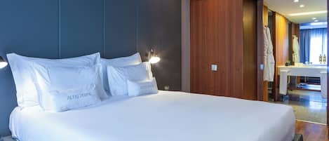 Un grand lit avec une literie blanche et des oreillers portant l’inscription « ALTIS PRIME », dans une chambre d’hôtel moderne avec des panneaux de bois et une salle de bain visible.