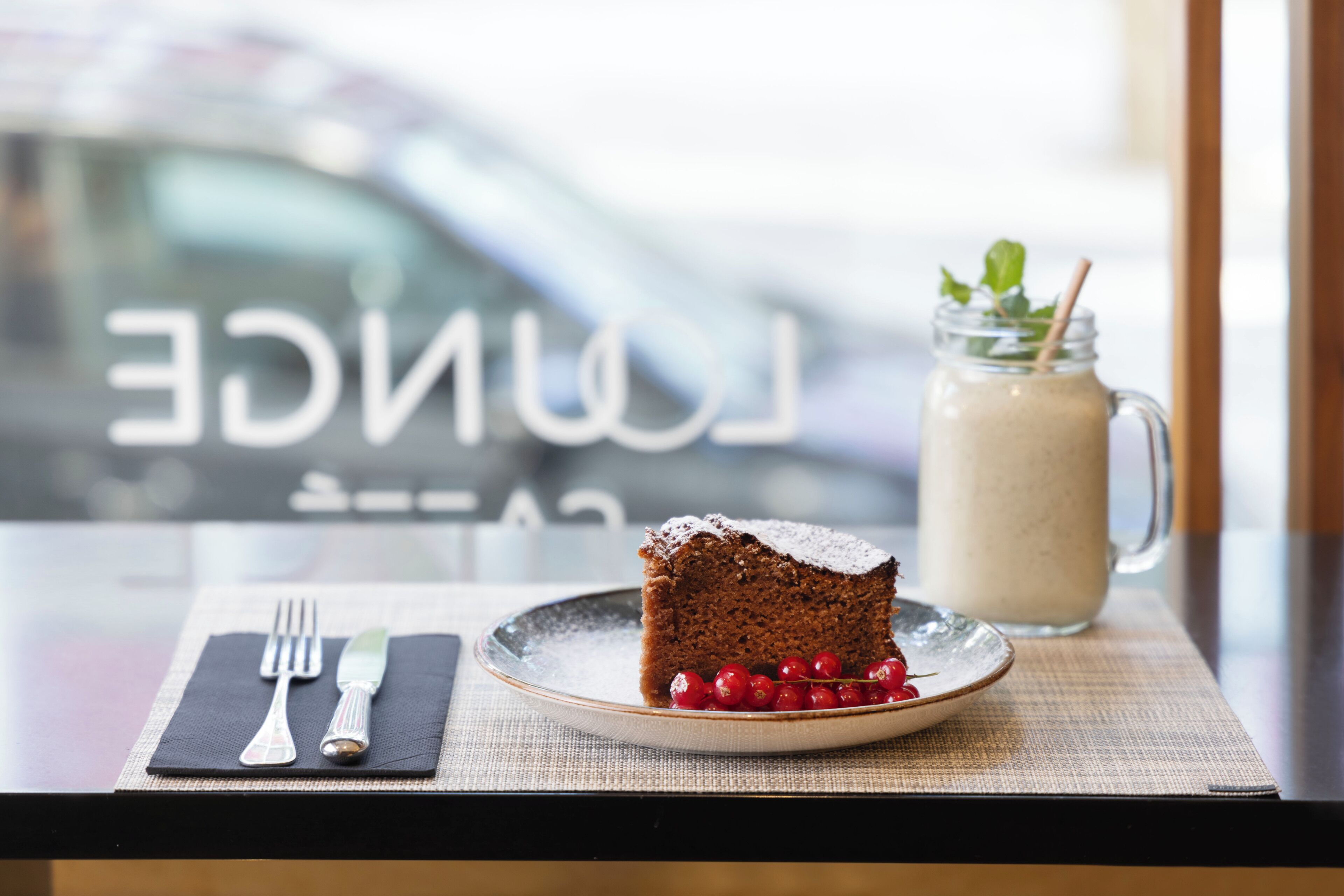 A slice of cake on a plate with a glass of milk, a fork, and a knife on a table.