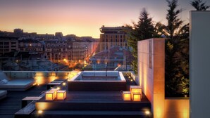 A rooftop terrace with seating areas, a pool, and a view of the city skyline at dusk.