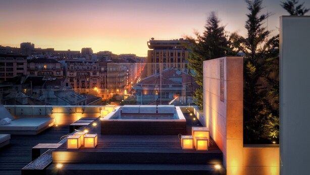 Une terrasse sur le toit, aménagée avec des espaces salon, une piscine et offrant une vue sur les toits de la ville au crépuscule.